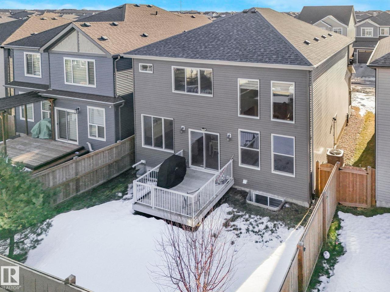 Rear exterior featuring a grey siding facade, multiple windows, a ground-level deck with white railing, and a fenced backyard - 8641 Mayday Wynd Sw, Edmonton, AB - Outdoor