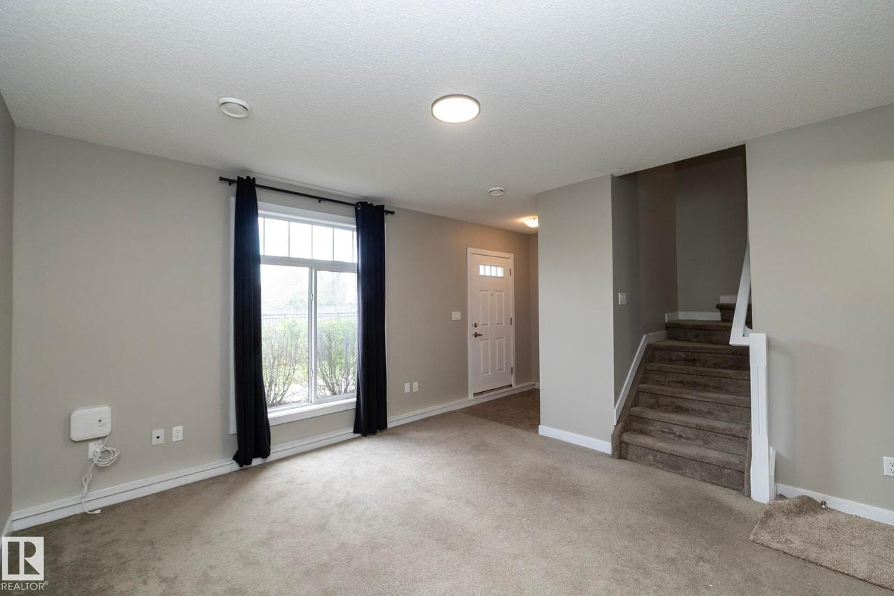 Open concept living space featuring neutral gray walls, plush carpet flooring, a multi-pane window with black drapery, and a visible staircase with carpeting - 117 Callaghan Drive Sw, Edmonton, AB - Indoor Photo Showing Other Room