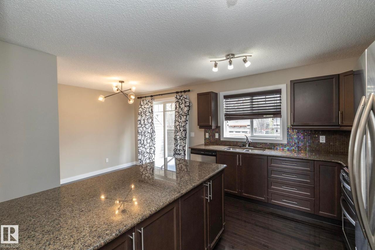 Kitchen featuring an expansive island with a granite countertop, dark wood-finish cabinetry, stainless steel appliances, a mosaic tile backsplash, and dark wood-finish flooring - 117 Callaghan Drive Sw, Edmonton, AB - Indoor Photo Showing Kitchen With Double Sink