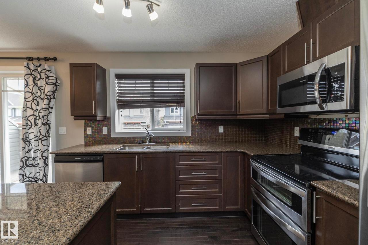Contemporary kitchen featuring dark wood cabinetry, granite-finish countertops, stainless steel appliances, a mosaic tile backsplash, and dark wood-finish flooring - 117 Callaghan Drive Sw, Edmonton, AB - Indoor Photo Showing Kitchen With Double Sink