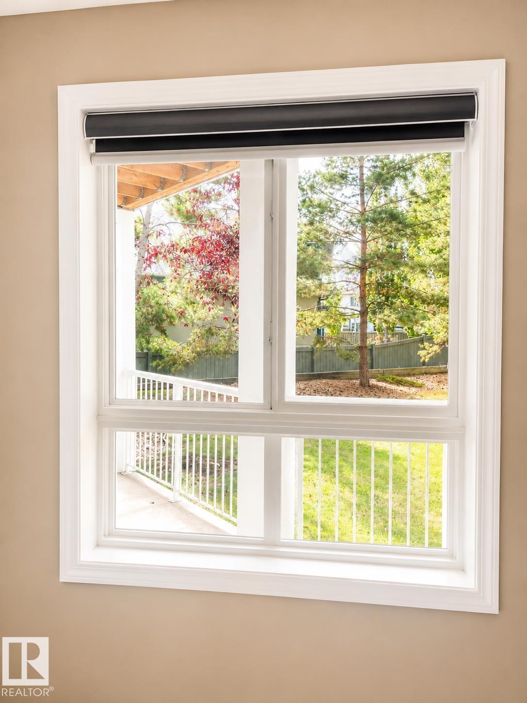 Double-paned window with bright white trim and integrated roller shades, offering a partial exterior patio deck with white railings and mature trees - #115 5340 199 St Nw, Edmonton, AB - Indoor Photo Showing Other Room