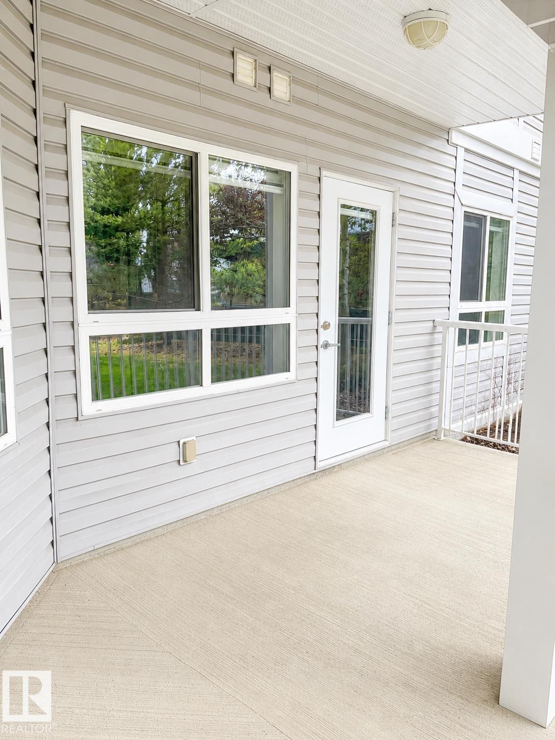 Covered concrete patio with textured finish, featuring light gray siding, multiple windows, a white framed door with glass paneling, and a white metal railing - #115 5340 199 St Nw, Edmonton, AB - Outdoor With Deck Patio Veranda With Exterior