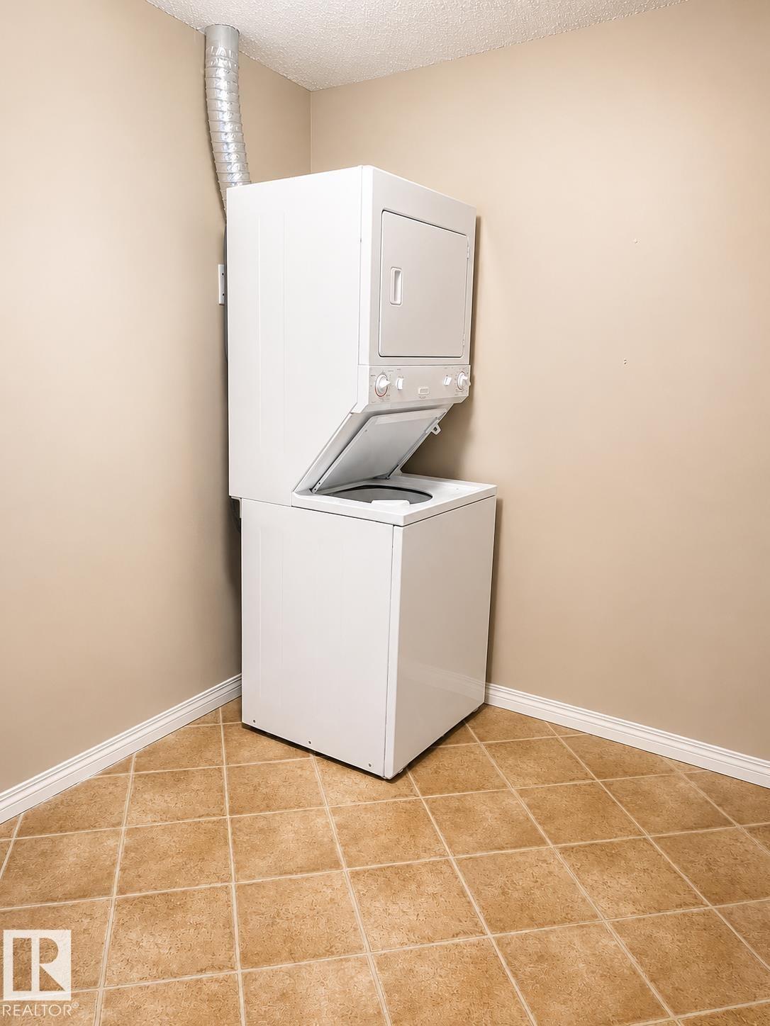 Dedicated laundry area featuring a stacked washer and dryer unit, neutral wall paint, white baseboards, and square tan floor tiles - #115 5340 199 St Nw, Edmonton, AB - Indoor Photo Showing Laundry Room