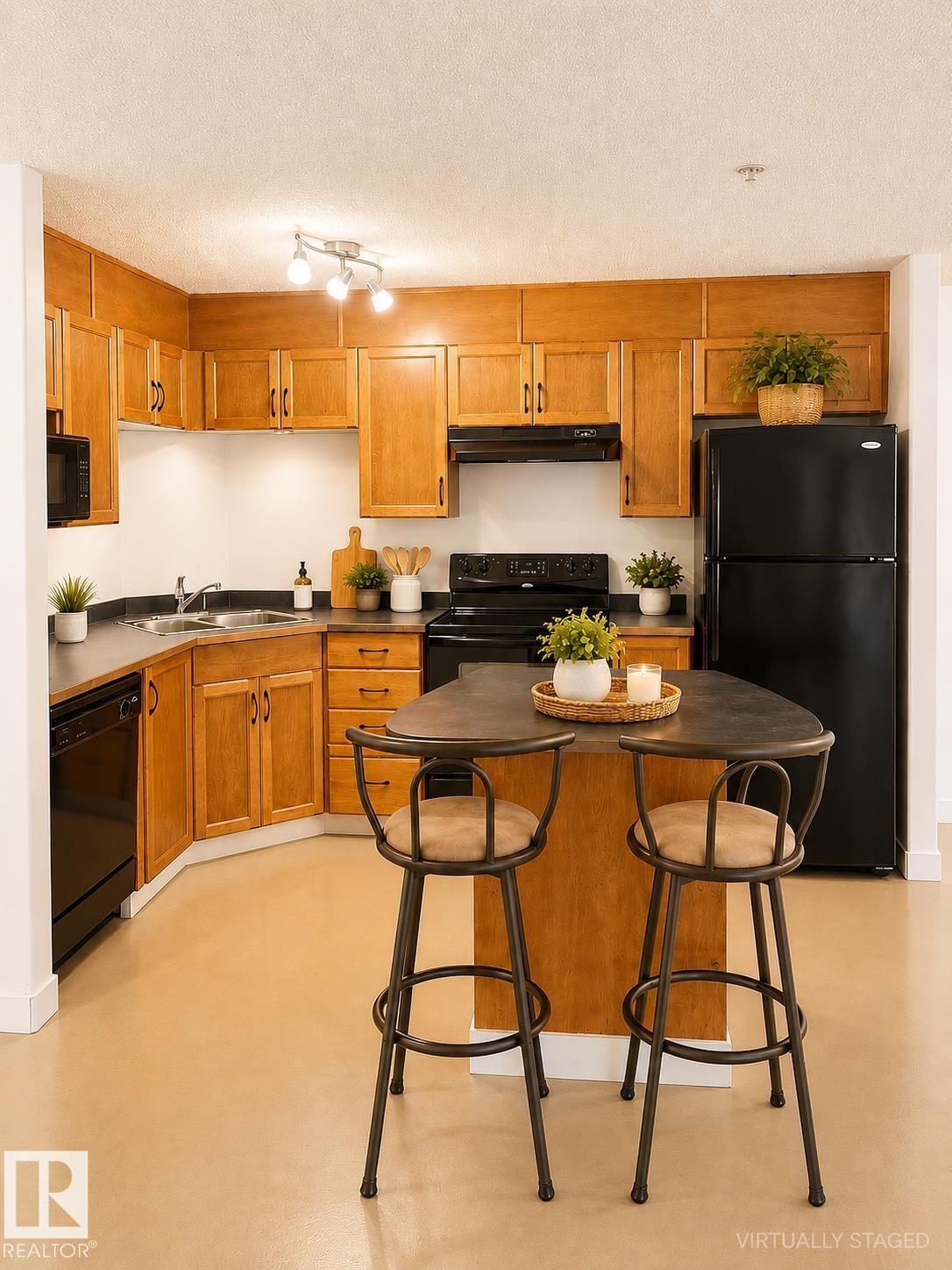 U-shaped kitchen with wood cabinetry, dark countertops, black appliances, and a central island with integrated seating - #115 5340 199 St Nw, Edmonton, AB - Indoor Photo Showing Kitchen With Double Sink