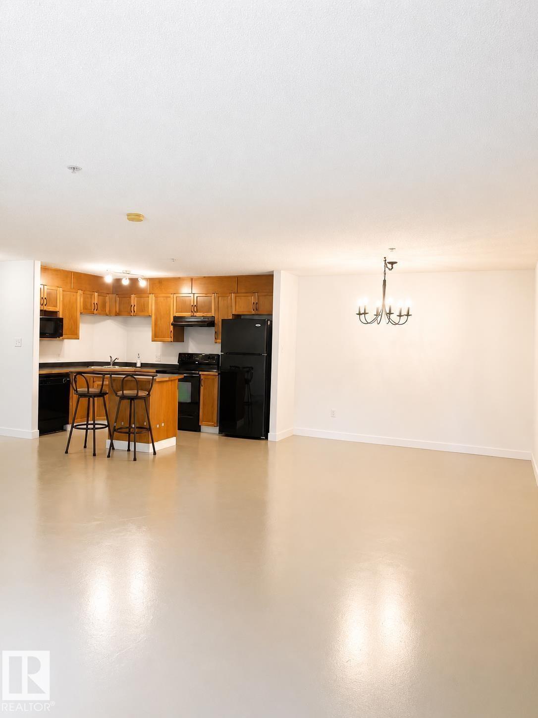 Open-concept kitchen and living area featuring a breakfast bar with seating, black appliances, warm wood cabinetry, and a black chandelier - #115 5340 199 St Nw, Edmonton, AB - Indoor Photo Showing Other Room