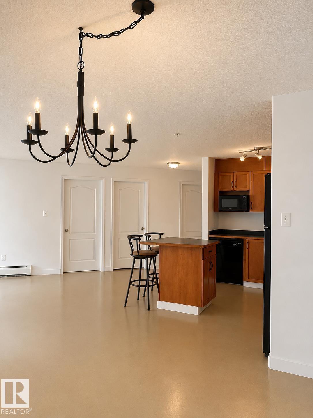 Spacious open-concept living area featuring a black chandelier, light-toned flooring, white walls, and a kitchen with wood-finish cabinetry, black countertops, and black appliances - #115 5340 199 St Nw, Edmonton, AB - Indoor