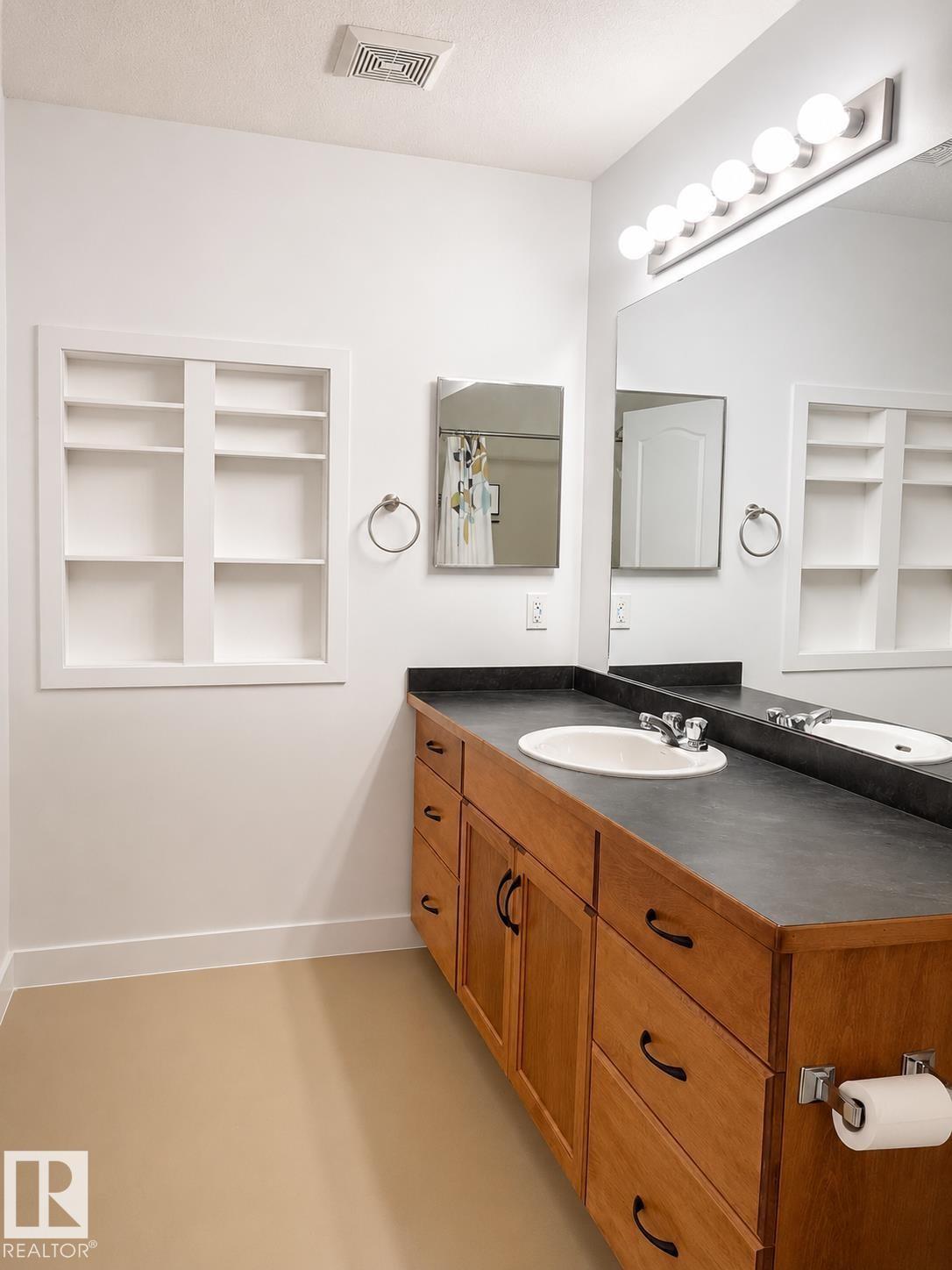 Bathroom featuring a wood-finish vanity with an integrated sink, dark countertop, and large mirror with globe lighting - #115 5340 199 St Nw, Edmonton, AB - Indoor Photo Showing Bathroom