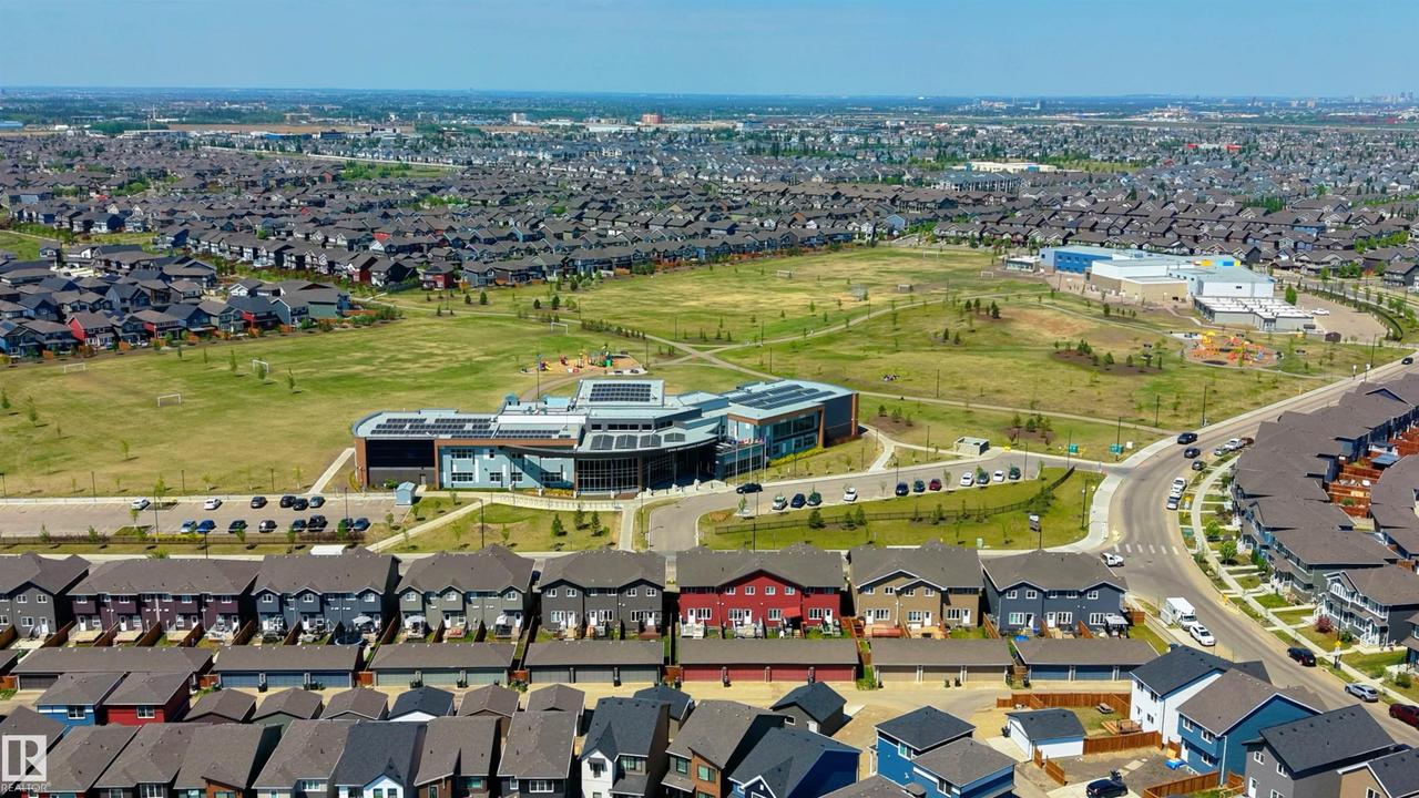 Expansive aerial perspective showcasing a residential development with numerous homes, a large central green space, a modern institutional building with solar panels, and curved roadway infrastructure - 2120 Muckleplum Crescent Sw, Edmonton, AB - Outdoor With View