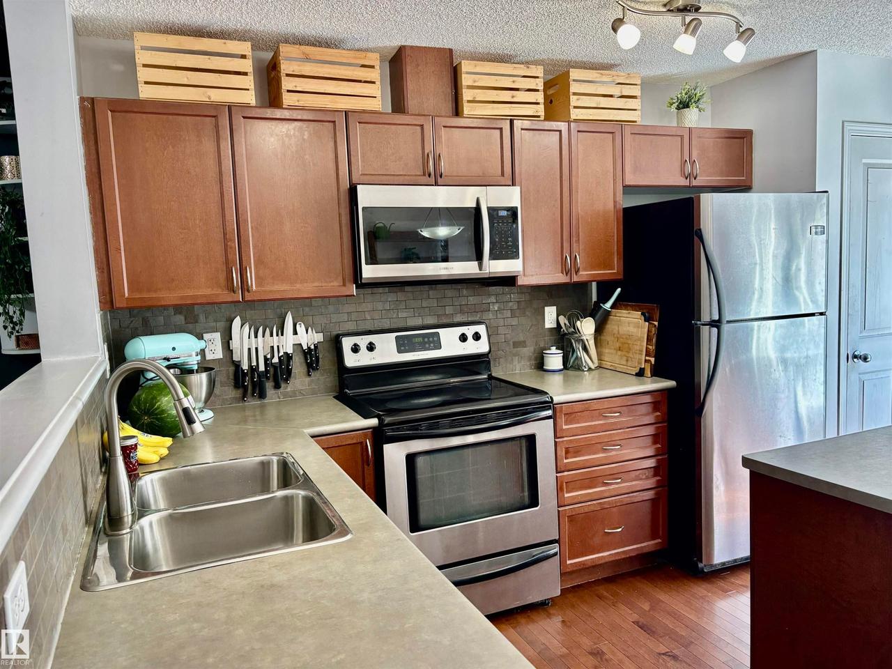 Kitchen featuring wood-finish cabinetry, stainless steel appliances, a grey mosaic tile backsplash, and light-toned countertops - #184 1804 70 St Sw, Edmonton, AB - Indoor Photo Showing Kitchen With Double Sink