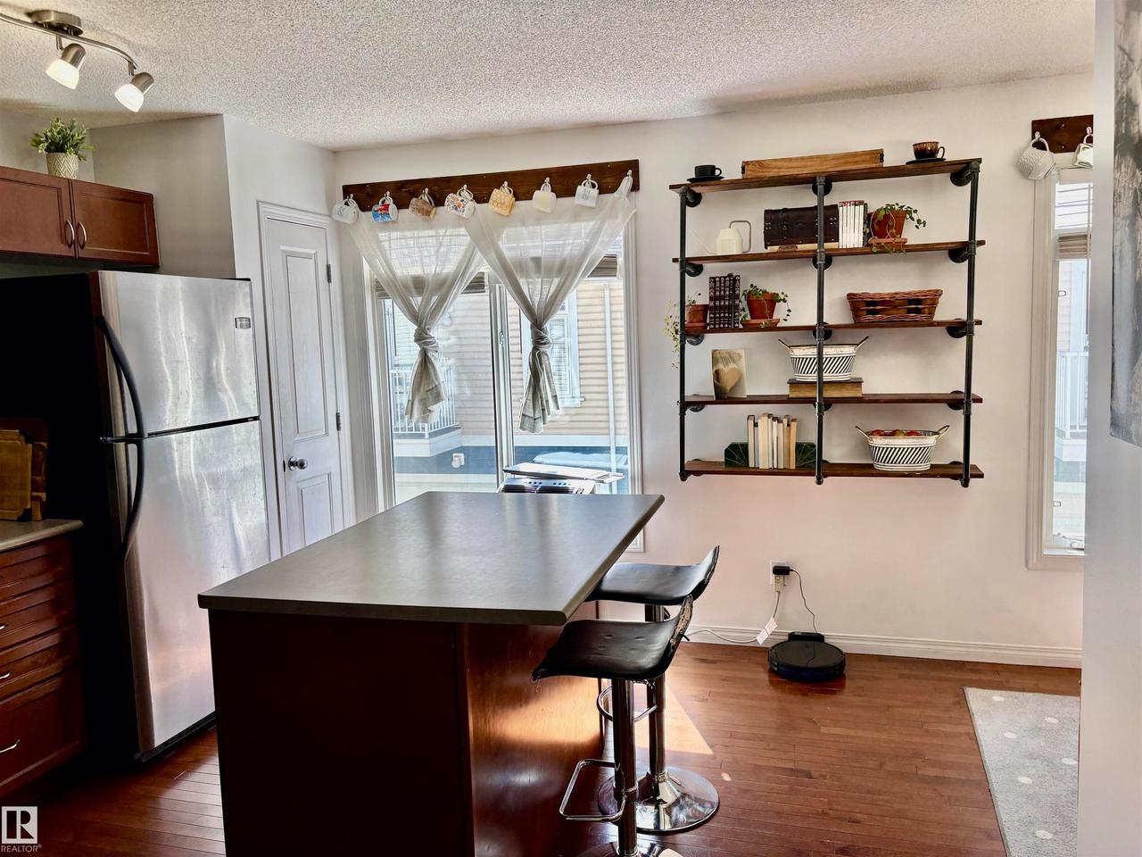 Kitchen area featuring an island with seating, stainless steel refrigerator, dark wood-finish flooring, and open shelving with metal pipe supports - #184 1804 70 St Sw, Edmonton, AB - Indoor