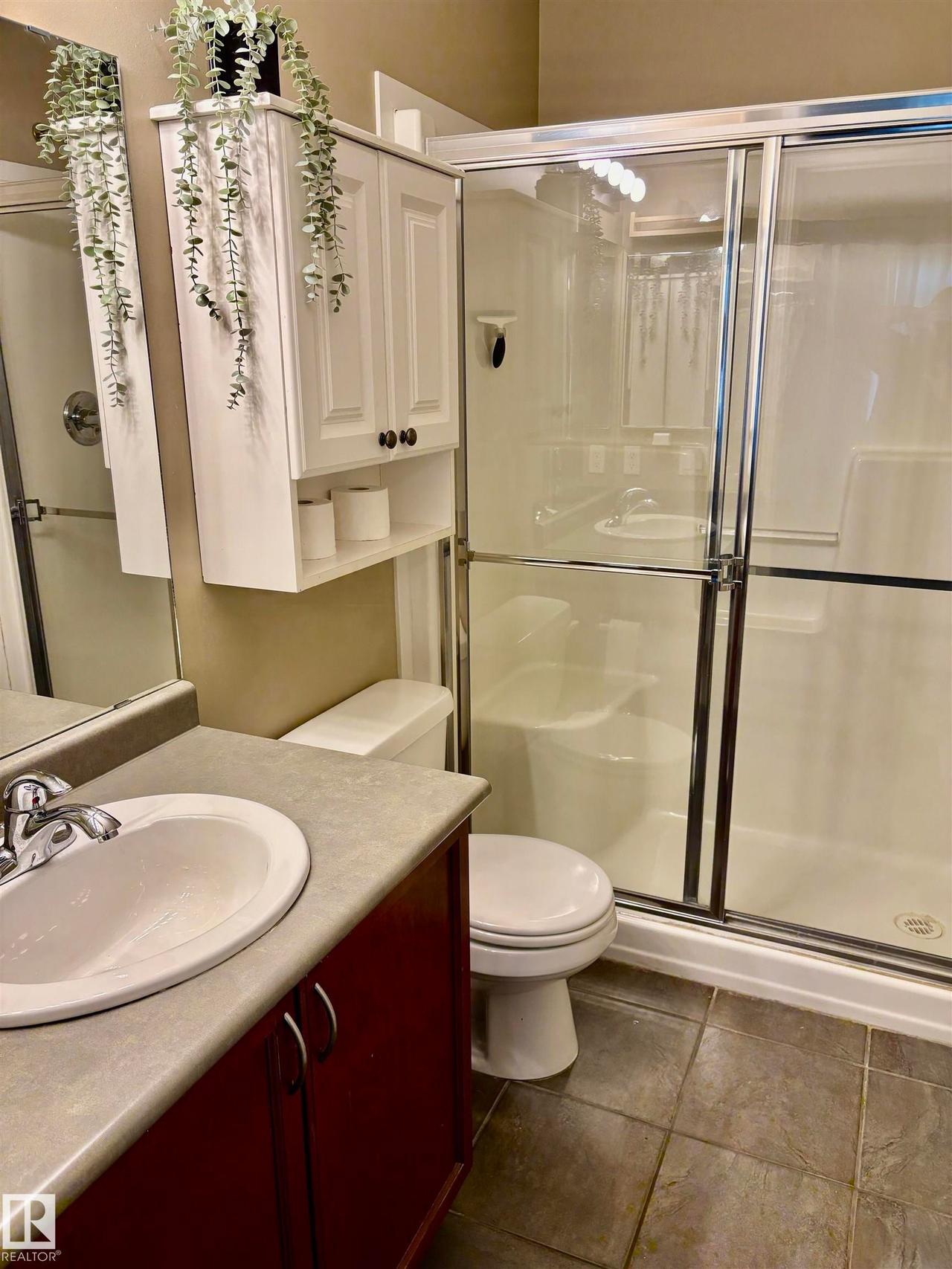 Three-piece bathroom featuring a shower with sliding glass doors, a vanity with a white basin and dark red cabinetry, and a wall-mounted storage cabinet - #184 1804 70 St Sw, Edmonton, AB - Indoor Photo Showing Bathroom