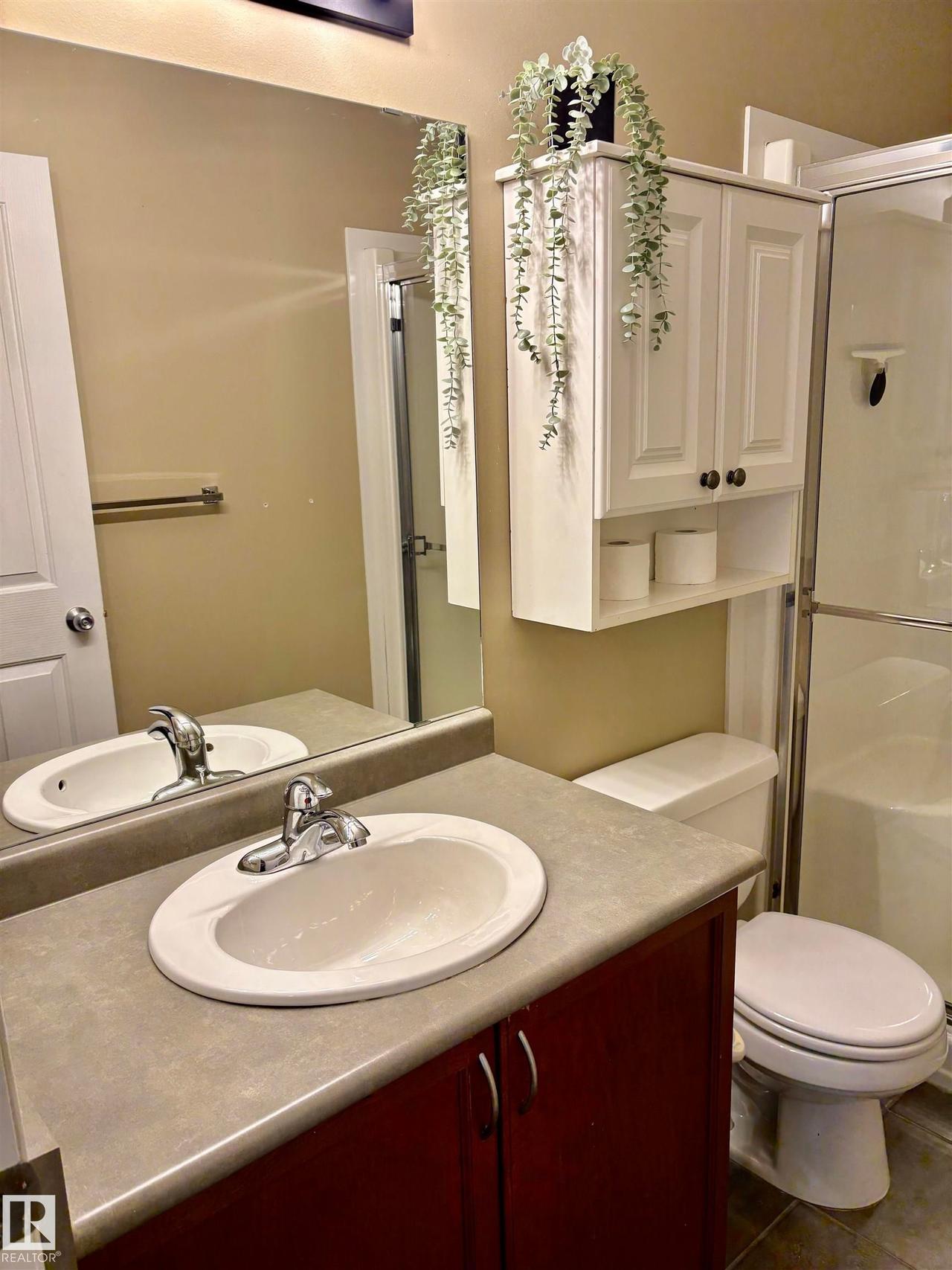 Bathroom featuring a vanity with a dark wood-finish cabinet and integrated sink, a large mirror, white wall-mounted cabinetry, a toilet, and a stand-up shower with a glass enclosure - #184 1804 70 St Sw, Edmonton, AB - Indoor Photo Showing Bathroom