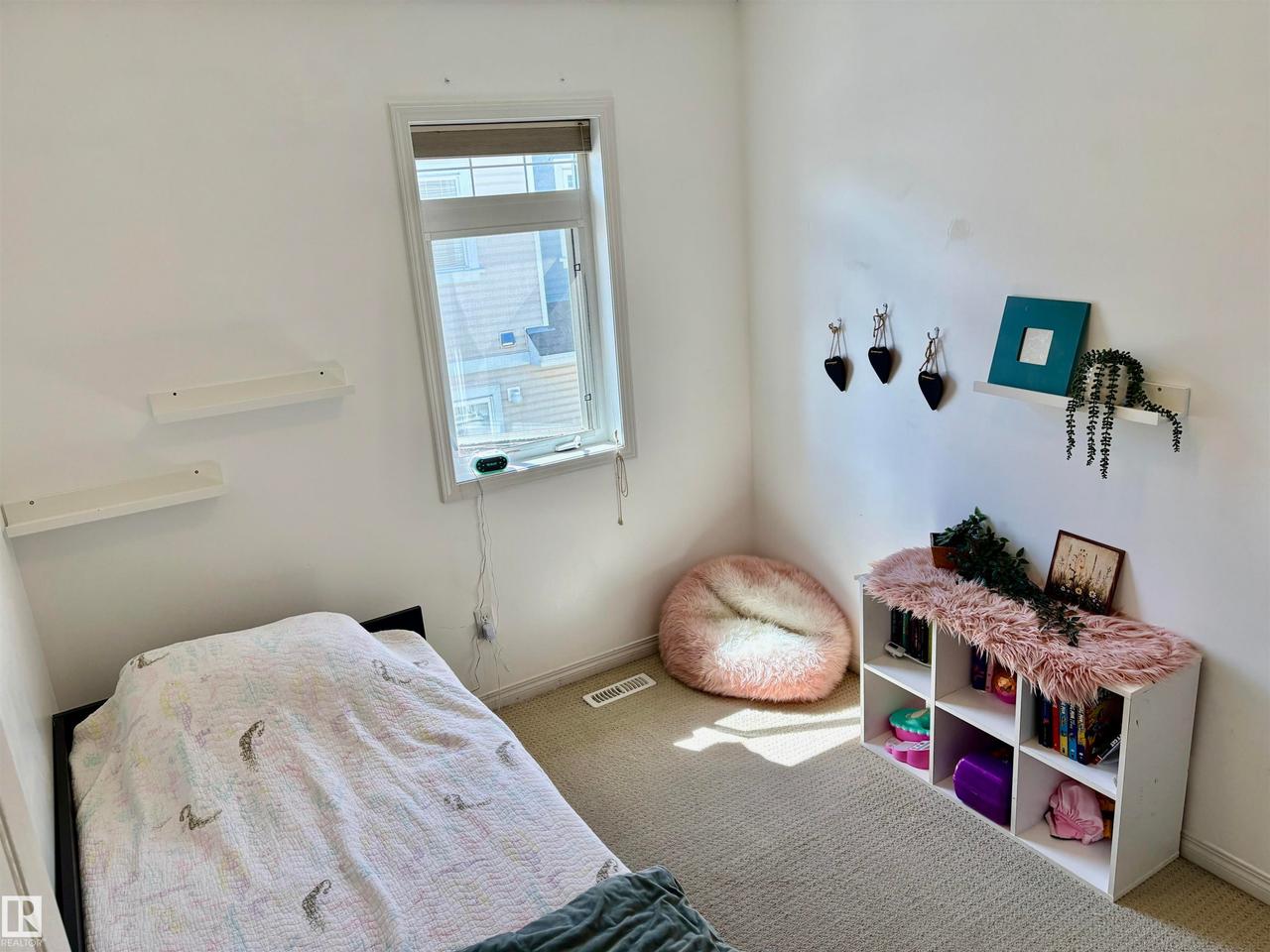 Bright room featuring a window with a roller shade, light-colored carpeting, white baseboards, and multiple white wall-mounted shelves - #184 1804 70 St Sw, Edmonton, AB - Indoor Photo Showing Bedroom