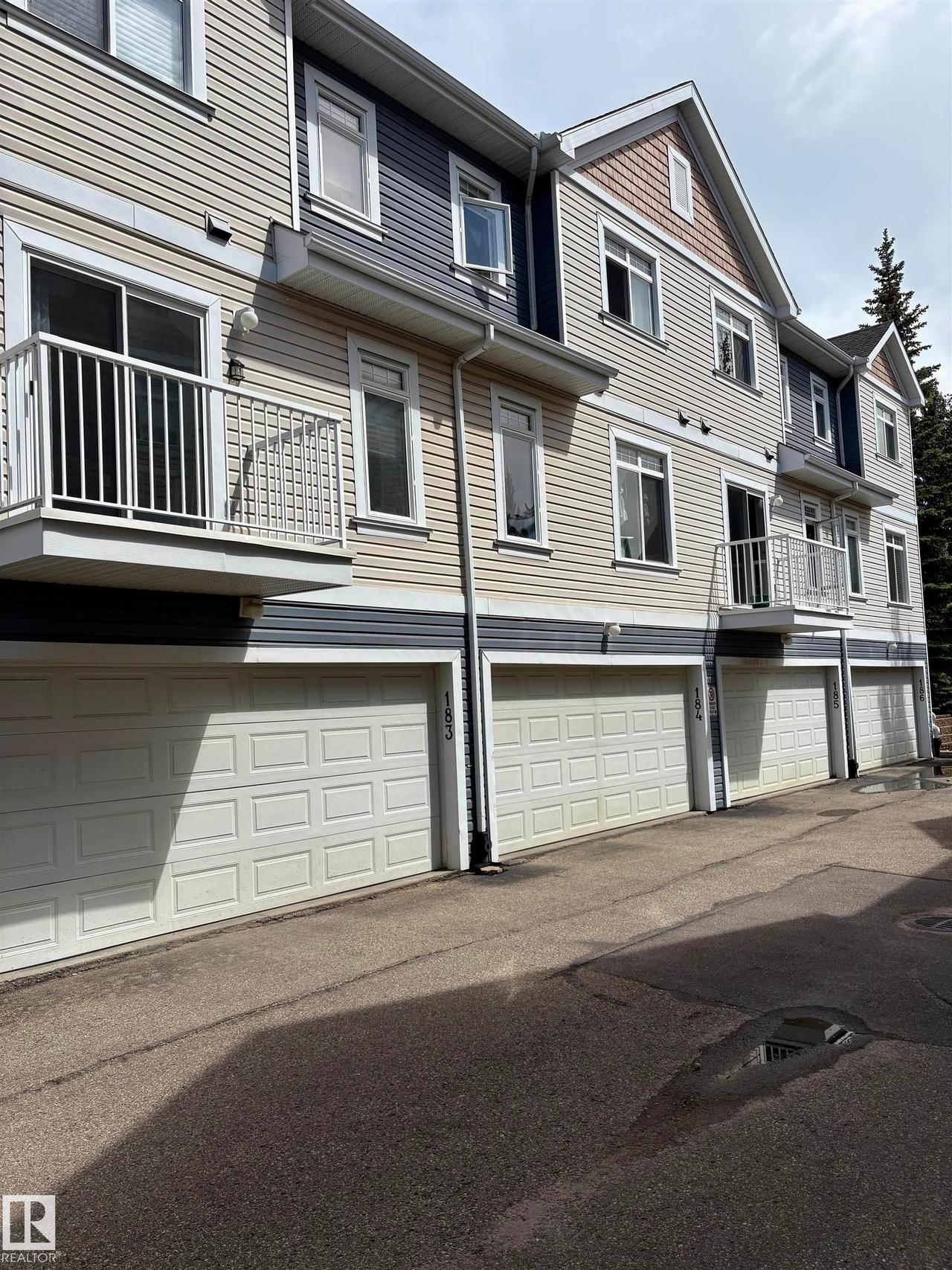 Multi-level exterior featuring light beige and dark blue horizontal siding, white trim, multiple balconies with white railings, and individual garage doors - #184 1804 70 St Sw, Edmonton, AB - Outdoor